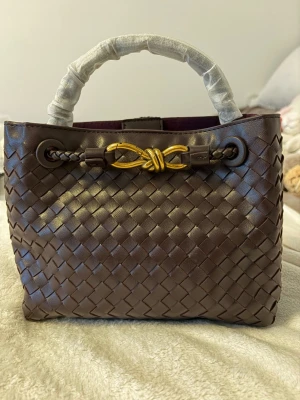 Bottega inspired bag - Mörkbrun handväska i flätat läder med guldfärgad metallknut framtill. Väskan har ett kort handtag, innerfack med dragkedja och magnetknäppning.