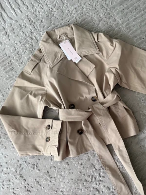 Kort beige trenchcoat från Gina Tricot  - Trendig kort trenchcoat i beige från Gina Tricot med klassisk dubbelknäppning, bred krage, axelklaffar och knytskärp i midjan. Jackan har dekorativa knappar vid ärmsluten och är tillverkad i ett slätt bomullsliknande material. Perfekt för en stilren look. Helt slut online och i butiker. 14+ collection från Gina Tricot! Passar även stl S