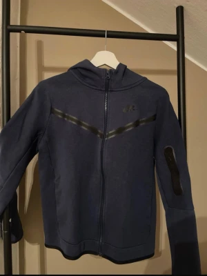 Mörkblå Nike hoodie med dragkedja - Snygg mörkblå hoodie från Nike med hel dragkedja framtill och huva. Har svarta detaljer i form av två diagonala ränder över bröstet och en ficka med dragkedja på ena ärmen. Tillverkad i mjukt material, perfekt för chill eller träning ( byxor ingår vid intresse!)