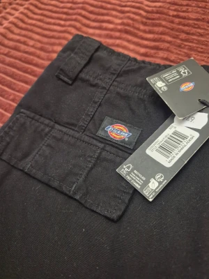 Vintage Svarta cargobyxor från Dickies W28 - Säljer ett par svarta cargobyxor från Dickies med raka ben och flera praktiska fickor på sidorna. Byxorna har klassisk cargomodell med knapp och dragkedja i midjan samt Dickies-logga på fickan. Perfekta för en avslappnad och street-inspirerad stil.