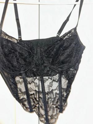 Svart spetskorsett med justerbara band - Säljer en snygg svart korsett med genomskinliga spetsdetaljer och justerbara axelband. Framsidan har vackert mönstrad spets och markerade sömmar för extra form. Baksidan är i mjukt tyg med spets nertill. Perfekt för dig som vill ha något extra till outfiten.