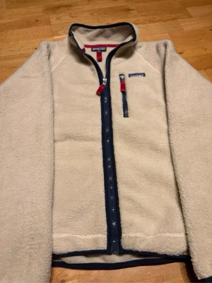 Beige teddyjacka från Patagonia - Säljer en beige teddyjacka från Patagonia med hel dragkedja i mörkblått och röda detaljer. Jackan har en bröstficka med dragkedja och stårkrage. Materialet är mjukt och fluffigt, perfekt för kyliga dagar. Klassisk och stilren design som funkar till det mesta.