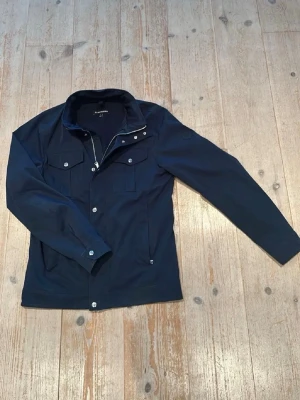 Bailey jacka från J. Lindeberg - Snygg marinblå vindjacka från J. Lindeberg med dragkedja och knappar framtill. Jackan har två bröstfickor med lock och två sidofickor. Långärmad modell i ett lätt och smidigt material, perfekt för vår och höst.