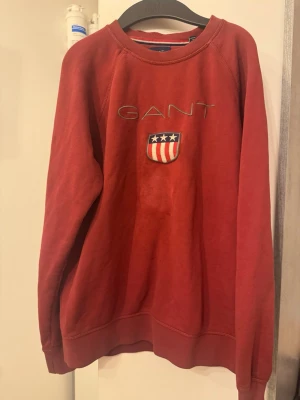 Röd sweatshirt från GANT - Röd sweatshirt från GANT med broderad logga och amerikanskinspirerat sköldmärke på bröstet. Tröjan har rund halsringning, ribbade muddar och klassisk passform. Perfekt för en avslappnad och stilren look.