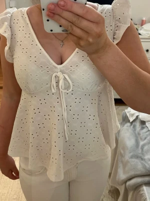 Vit blus med spets och knyt - Söt vit blus med broderade spetsdetaljer och v-ringning. Toppen har korta volangärmar och ett dekorativt rosett knyt framtill. Luftig och feminin passform, perfekt för varma dagar.