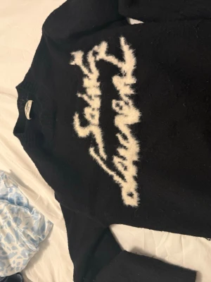 Svart stickad tröja Saint Laurent - Cool svart stickad tröja från Saint Laurent med ett vitt, fluffigt grafiskt mönster över bröstet. Tröjan har ribbstickad krage, ärmslut och nederkant. Perfekt för dig som gillar streetwear och vill ha något unikt i garderoben.
