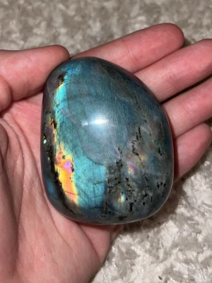 Polerad labradorit sten - Unik polerad labradorit med skimrande blå, gröna och gyllene nyanser. Stenen har en mjukt rundad form och vackra naturliga mönster som reflekterar ljuset på ett magiskt sätt. Perfekt som dekoration eller för smyckestillverkning.