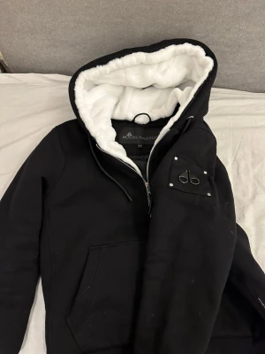 Svart pufferjacka Moose Knuckles - Snygg svart pufferjacka från Moose Knuckles med stor huva fodrad i vit päls. Jackan har dragkedja, quiltat innerfoder och två fickor framtill. Ikonisk logga på ärmen och detaljer i metall. Perfekt för vädret just nu! Tagsen medföljer.