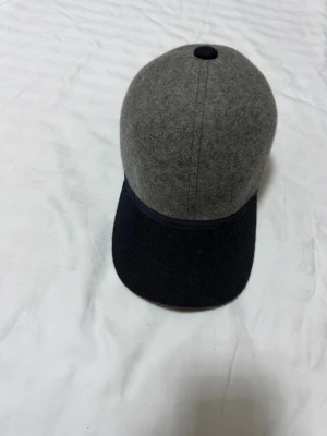 Grå och svart keps i ullmix - Snygg keps med grå ovandel och svart skärm, tillverkad i mjuk ullmix som ger en stilren och modern look. Klassisk form med knapp på toppen och böjd skärm. Perfekt för dig som vill ha en enkel men trendig accessoar.