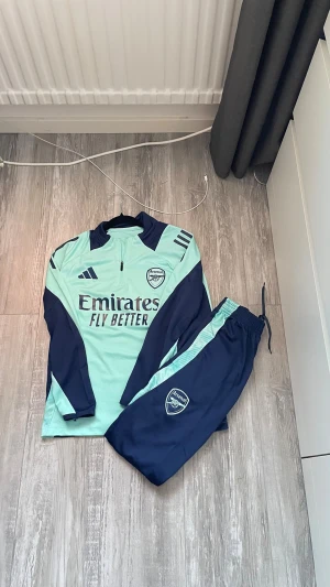Arsenal träningsset från Adidas - Snyggt Arsenal träningsset från Adidas i ljusblått och marinblått. Setet består av en långärmad tröja med halv dragkedja och matchande byxor med dragsko. Både tröjan och byxorna har Arsenal-logga och Adidas-detaljer. Perfekt för fotbollsträning eller chill. Storlek: 16YR - passar för 12-16 åringar