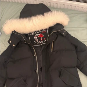 Svart dunjacka från Moose Knuckles - Säljer en svart dunjacka från Moose Knuckles med stor luva och fluffig vit fuskpäls. Jackan har silverfärgade detaljer, dragkedja och tryckknappar samt logga på ärmen. Perfekt för kalla vinterdagar och riktigt snygg streetstil.