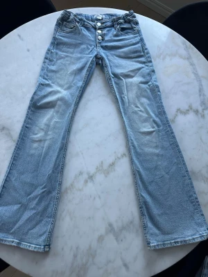 Ljusblå bootcut jeans med knappar - Säljer ett par ljusblå bootcut jeans med tre synliga knappar framtill. Jeansen har en lätt tvättad look och utsvängda ben. Perfekta för dig som gillar retrostil och vill ha ett par bekväma jeans med coola detaljer.