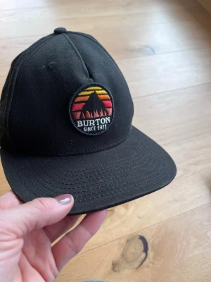 Svart keps från Burton - Snygg svart keps från Burton med rund patch framtill som har ett bergmotiv i rött, orange och gult samt texten 'Burton Since 1977'. Klassisk form med böjd skärm och justerbar baksida. Perfekt för en avslappnad streetwear-look.