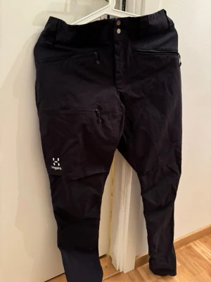 Svarta byxor från Haglöfs med dragkedjor - Säljer ett par svarta byxor från Haglöfs med flera praktiska dragkedjefickor framtill. Byxorna har en modern, tapered passform och är tillverkade i slitstarkt syntetmaterial. Perfekta för dig som gillar funktionella och stilrena plagg.