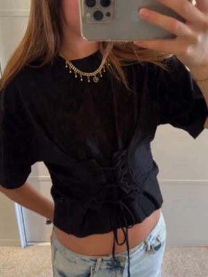 Svart t-shirt med snörning - Snygg svart croppad topp med snörning framtill som ger en edgy vibe. Toppen har korta ärmar och en figurnära passform som framhäver midjan. Perfekt att matcha med jeans eller kjol för en trendig look.