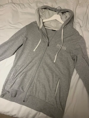 Grå zip hoodie från Hugo Boss - Snygg ljusgrå hoodie från Hugo Boss med dragkedja framtill och vit logga på bröstet. Hoodien har vita dragsnören, fickor på sidorna och ribbade muddar. Perfekt för en chill och stilren look. Mjuk bomullsblandning för extra komfort.