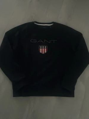 Svart sweatshirt från GANT - Snygg svart sweatshirt från GANT med klassisk logga och en broderad logga på bröstet. Tröjan har rund halsringning och en stilren design som passar perfekt till jeans eller chinos.
