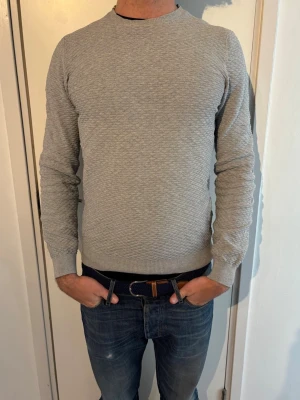 Massimo Dutti tröja  - Säljer en ljusgrå tröja från Massimo Dutti med diskret strukturmönster. Tröjan har rund halsringning, långa ärmar och ribbade muddar vid ärmslut och nederkant. Perfekt till jeans eller chinos för en clean look.