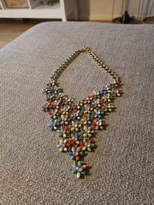Färgglatt statementhalsband med pärlor - Säljer ett statementhalsband med guldfärgad kedja och massor av små pärlor i form av blommor i blått, gult, orange, grönt och rött. Halsbandet har en unik och iögonfallande design som verkligen sticker ut. Perfekt för dig som vill ha något färgglatt och lekfullt.  En av stenarna skiner inte lika mycket längre. Men tycker inte det gör något. Se skick på bilderna. Strass! 