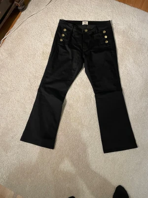 Svarta bootcut jeans från Isay - Snygga svarta bootcut jeans från Isay med hög midja och dekorativa guldknappar framtill. Jeansen har klassiska fickor både fram och bak och är tillverkade i ett mjukt material av bomull, polyester och elastisk som ger en skön passform.