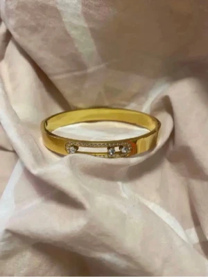 Guldigt armband med gnistrande stenar - Snyggt armband i guldfärgad metall med en unik säkerhetsnål-design framtill, dekorerad med små gnistrande stenar som ger extra bling. Armbandet har en stilren och modern look och öppnas enkelt med ett gångjärn. Perfekt för dig som gillar trendiga accessoarer.