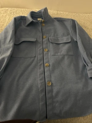 Ljusblå overshirt med fickor - Snygg ljusblå overshirt med klassisk krage och stora bröstfickor med lock. Stängs med knappar framtill och har knappar vid ärmsluten. Overshirten har en rak passform och är tillverkad i ett mjukt, filtliknande material.