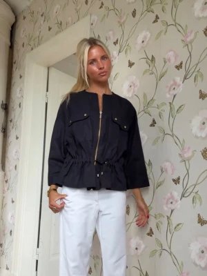 Svart jacka med dragkedja från Zara  - Stilren svart jacka från Zara med dragkedja framtill, två bröstfickor med knappar och justerbar midja med dragsko. Jackan har rund halsringning och raka ärmar. Materialet ser ut att vara en lättare bomullsblandning, perfekt för lager på lager. Ny med prislappar!