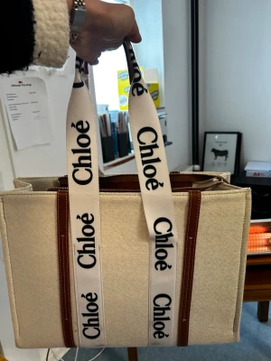 Chloé Woody beige handväska - Trendig beige handväska från Chloé i canvas med bruna läderdetaljer och breda vita axelband med svart Chloé-logga. Väskan har en boxig form och rymligt huvudfack, perfekt för att bära med sig allt man behöver. Snygg och stilren design som sticker ut.