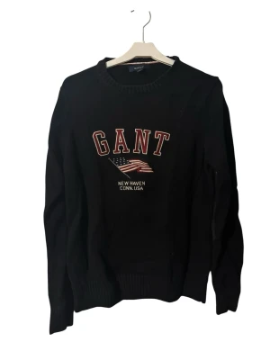 Svart stickad tröja från GANT - Snygg svart stickad tröja från GANT med broderad logga och amerikansk flagga på bröstet. Rund halsringning och ribbade muddar vid ärmslut och nederkant. Perfekt för dig som gillar klassisk college-stil.