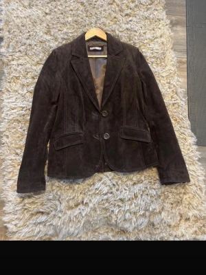 Vintage Mörkbrun kavaj i mocka - Snygg vintage mörkbrun kavaj från CUE i mjuk mocka med två knappar framtill och klassiska fickor med lock. Kavajen har en figurnära passform och helfodrad insida i blankt tyg. Perfekt för dig som vill ha en stilren och trendig look.