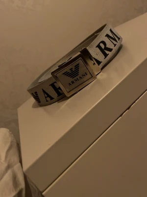 Grått Armani bälte med silverdetaljer - Snyggt grått bälte från Armani med tydlig logga och text längs hela bandet. Bältet har ett rektangulärt spänne i silverfärg med Armani-loggan framtill. Perfekt accessoar för att lyfta din outfit.