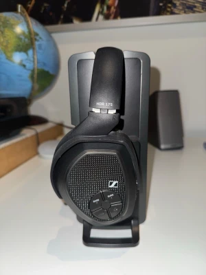 Sennheiser HDR 175 trådlösa hörlurar - Snygga svarta over-ear hörlurar från Sennheiser, modell HDR 175. De har en modern design med mjuka öronkuddar i konstläder och sitter på en laddningsstation. Hörlurarna har inbyggda knappar för volym, bass och surround direkt på kåpan.