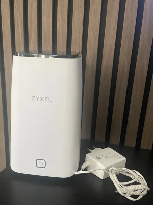Zyxel NR5103E 5G Router | mobilt bredband simkort |från operatörer tre - Säljer en stilren vit router från Zyxel med svart topp och tillhörande strömadapter. Routern har en modern, rektangulär design med rundade hörn och en knapp på framsidan. Perfekt för dig som vill ha stabilt internet hemma.