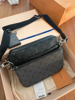 Louis Vuitton Monogram axelväska - Snygg axelväska från Louis Vuitton i svart och grått med klassiskt monogrammönster. Väskan har justerbar axelrem i textil, två fack med dragkedja och en liten avtagbar pouch. Tillverkad i slitstarkt canvasmaterial med silverfärgade metalldetaljer.