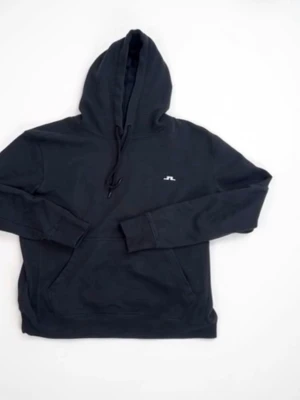 Svart hoodie från J.Lindeberg - Snygg svart hoodie från J.Lindeberg med liten vit logga på bröstet. Klassisk modell med huva och dragsko samt stor magficka framtill. Tillverkad i mjukt bomullsmaterial som är skönt att bära. Perfekt för en chill och stilren look.