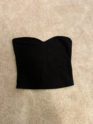 Svart axelbandslös tubtopp - Säljer en svart axelbandslös tubtopp med lätt hjärtformad urringning. Toppen är croppad och har en stilren look som passar till många outfits. Materialet känns mjukt och har en lätt struktur. Perfekt för dig som vill ha en enkel men snygg topp.