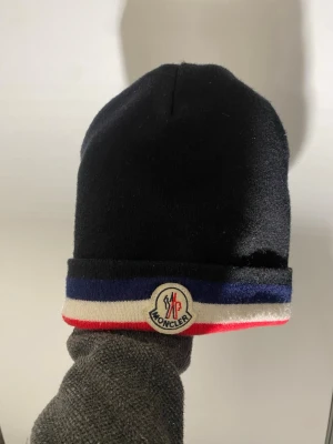 Svart Moncler mössa - Snygg svart mössa från Moncler med uppvikt kant i blått, vitt och rött. Framtill sitter Monclers klassiska logga. Perfekt accessoar för dig som vill ha en stilren och trendig look. Mössan är mjuk och skön, troligen i ull- eller bomullsblandning.