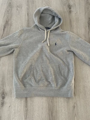 Grå hoodie från Polo Ralph Lauren - Snygg grå hoodie från Polo Ralph Lauren med klassisk huva och vita dragsnören. Tröjan har en känguruficka framtill och den ikoniska lilla loggan broderad på bröstet. Tillverkad i mjukt bomullsmaterial som är perfekt för chill dagar.
