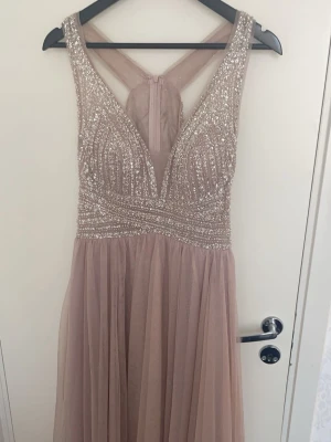 Glittrig långklänning i rosa champagne - Säljer en elegant långklänning i rosa champagnefärg med glittrig överdel och meshdetaljer. Klänningen har v-ringning, breda axelband och ett flytande, skirt kjolparti. Perfekt för bal eller festliga tillfällen. Ingen synlig logga på märket.
