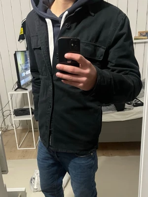 Svart overshirt med fickor - Säljer en svart overshirt med knappstängning och två bröstfickor. Overshirten har en klassisk krage och är tillverkad i ett kraftigare bomullstyg som är garment dyed för en schysst tvättad look. Perfekt att slänga över en hoodie för en avslappnad stil.