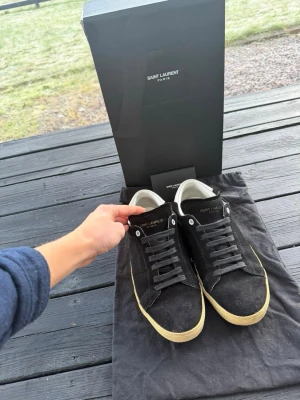 Saint Laurent svarta sneakers i mocka - Svarta Saint Laurent sneakers i mocka med vita detaljer och beige sula. Klassisk låg modell med svarta skosnören och diskret logga på plösen. Snygg och stilren design som passar till det mesta. Levereras med originalkartong och dustbag. Äktehetsbevis finns!