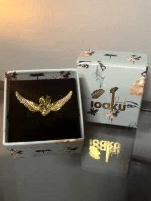 Guldfärgad ring med vingar från Ioaku - Unik guldfärgad ring från Ioaku med detaljerat motiv av ett hjärta och stora utbredda vingar. Ringen levereras i original ask. Perfekt för dig som gillar statement-smycken och vill sticka ut. Reglerbar ring, Finns inte att köpa i längre i butik. 