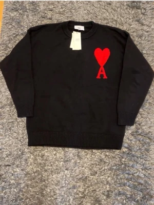 Svart stickad tröja från Ami Paris - Svart stickad tröja från Ami Paris med rund hals och ribbade muddar. På bröstet finns ett stort rött hjärta med bokstaven A, vilket är Ami Paris ikoniska logga. Tröjan har en klassisk passform och är perfekt för dig som gillar stilrena detaljer.