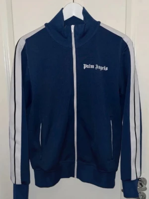 Blå zip hoodie från Palm Angels - Snygg blå zip hoodie från Palm Angels med vita ränder längs ärmarna och vit dragkedja. Klassisk streetwear-look med hög krage och logga på bröstet. Tillverkad i mjukt och bekvämt material, perfekt för en chill och trendig stil.