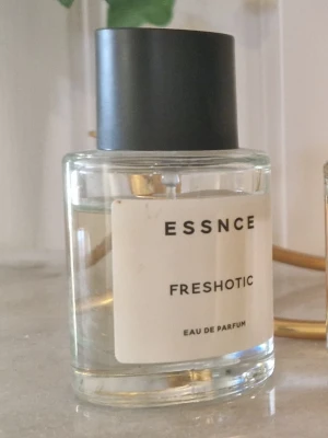 ESSNCE Freshotic Eau de Parfum - Minimalistisk parfymflaska från ESSNCE med doften Freshotic. Flaskan är cylindrisk i genomskinligt glas med svart rund kork och vit etikett med svart text. Modern och stilren design som passar perfekt på parfymhyllan.