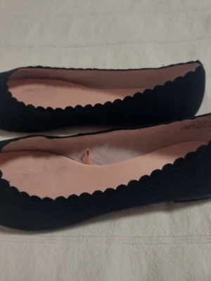Svarta ballerinaskor med vågig kant - Säljer ett par svarta ballerinaskor från H&M med snygg vågig kant runt öppningen. Skorna har en klassisk rundad tå och är gjorda i ett mjukt tygmaterial med ljusrosa innersula. Perfekta för en stilren och enkel look. Har bara provats men det rosa tyget invändigt är lite gråflammigt.