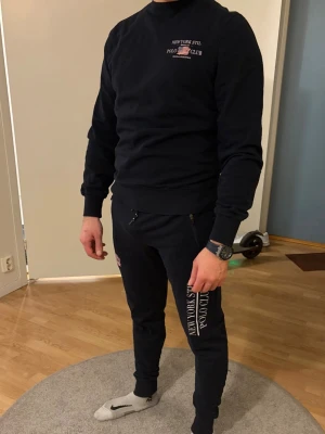 mjukis set New York Stil Polo Club - Säljer ett par svarta mjukisbyxor från New York Stil Polo Club med vit logga och text längs benet. Byxorna har dragsko i midjan och smal passform med muddar vid bensluten. Perfekta för chill eller träning.