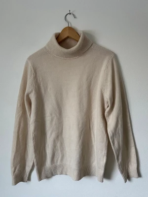 Beige Uniqlo polotröja i ullmix - Klassisk beige Uniqlo stickad polotröja med ribbade muddar och hög krage. Tröjan har en enkel och stilren design, perfekt för lager på lager. Materialet ser ut att vara en mjuk ull- eller ullmix som håller dig varm under kyliga dagar. 8A7