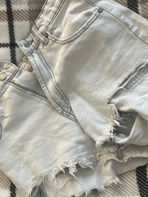 Ljusa denimshorts med fransar från Zara - Säljer ett par ljusa denimshorts från Zara i storlek 34. Shortsen har råa, fransiga kanter och klassisk femficksmodell. Perfekta för sommaren och har en avslappnad vibe med slitna detaljer. Passar dig som gillar en trendig och chill stil.
