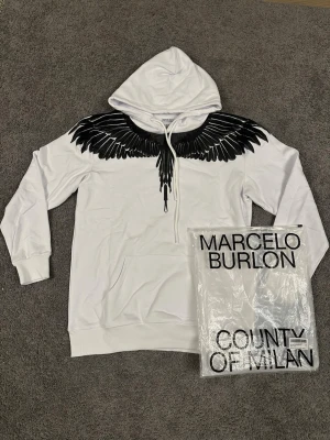 Vit hoodie Marcelo Burlon med vingar - Snygg vit hoodie från Marcelo Burlon med ikoniskt svart vingtryck över axlar och bröst. Klassisk känguruficka framtill, ribbade muddar och huva med dragsko. Perfekt statement-plagg för dig som vill sticka ut med streetwear-stil.